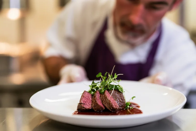 Chef plating sliced medium-rare steak with sauce and microgreens on white plate in restaurant kitchen