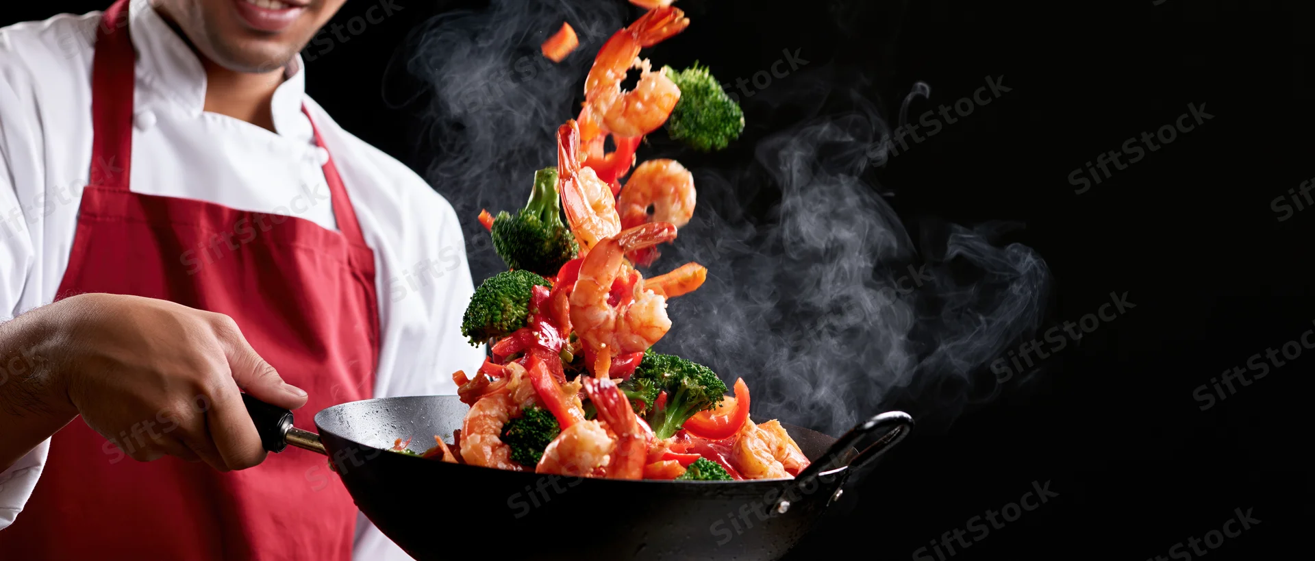 Chef tossing shrimp stir-fry in a wok with broccoli and steam against black background