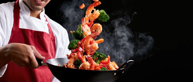 Chef tossing shrimp stir-fry in a wok with broccoli and steam against black background