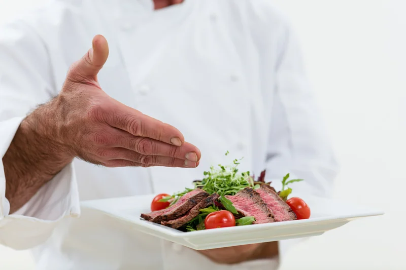 Chef presenting a plated steak salad with tomatoes and greens on a white plate