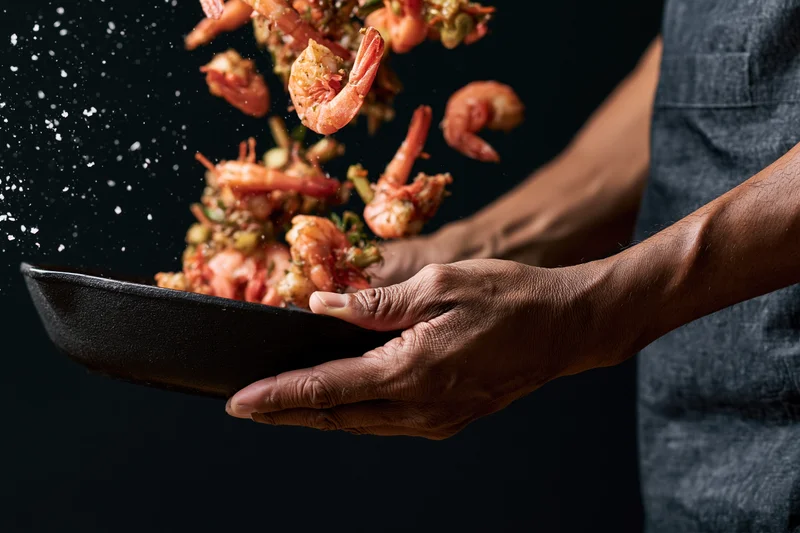 Chef tossing shrimp and vegetables midair above a dark skillet
