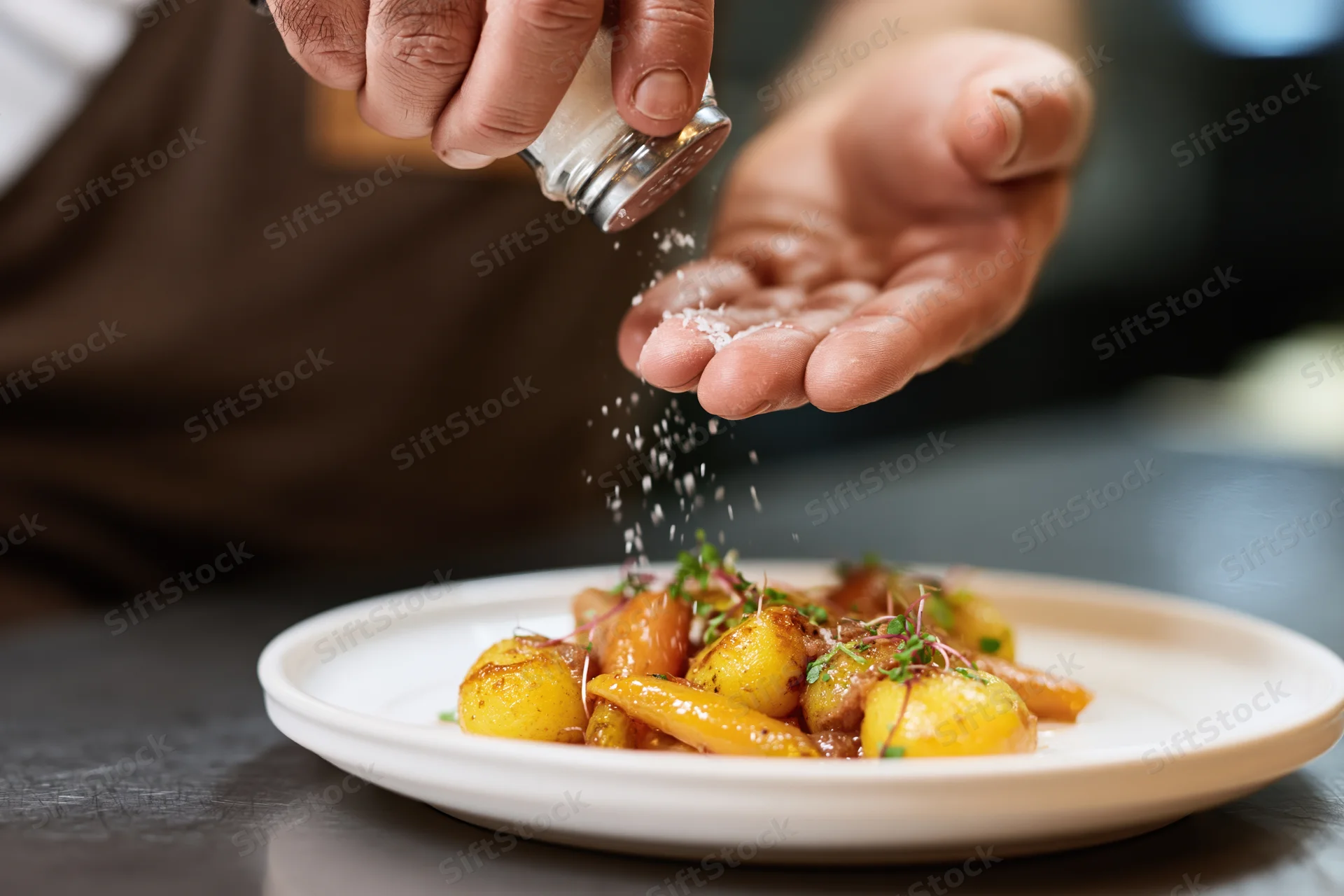 Chef's hands sprinkling salt from a glass shaker over a plated dish of roasted potatoes and vegetables