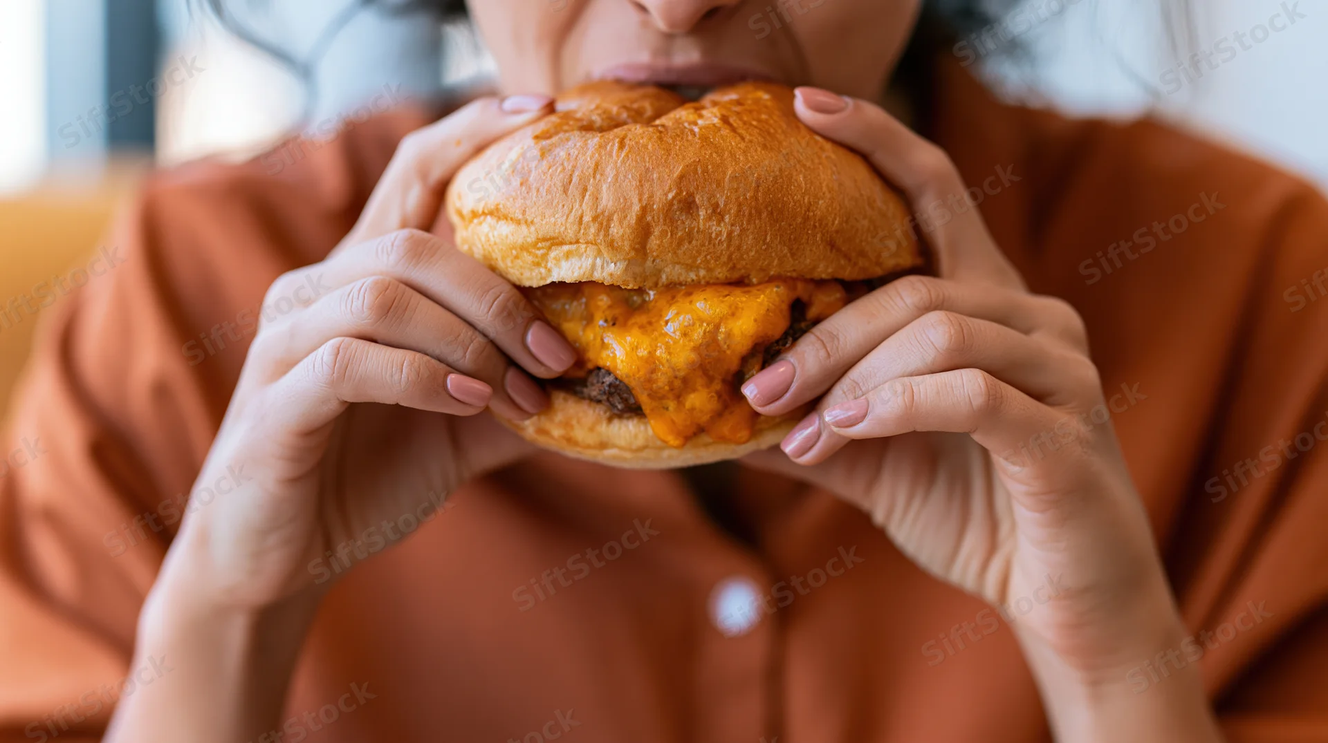 Woman biting into a thick cheeseburger with melted cheese, holding it with both hands, indoors