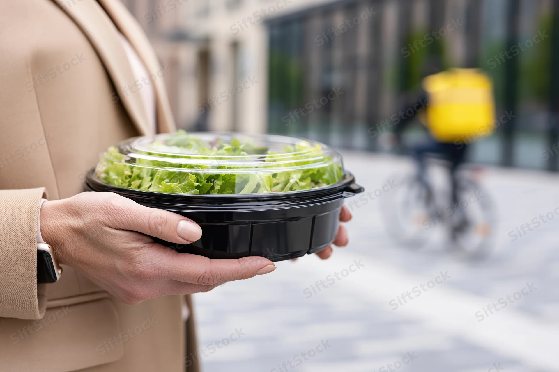Woman holding a black plastic takeout container with salad on a city sidewalk with a delivery bicycle in the background