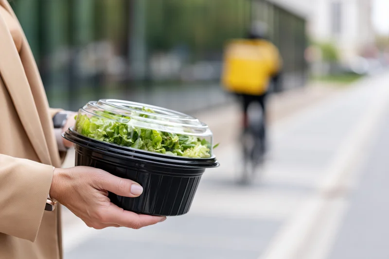 Woman holding a black plastic takeout container with salad on a city street with a delivery rider on bicycle in background