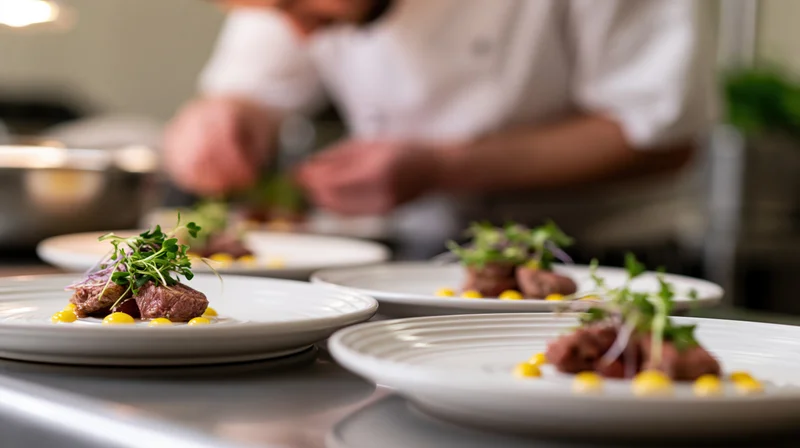 Fine dining dishes plated with steak and microgreens on stainless steel counter with chef in background