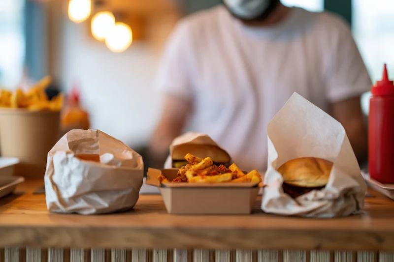 Two burgers wrapped in parchment paper on a wooden table with fries and ketchup bottle, man in background at restaurant counter