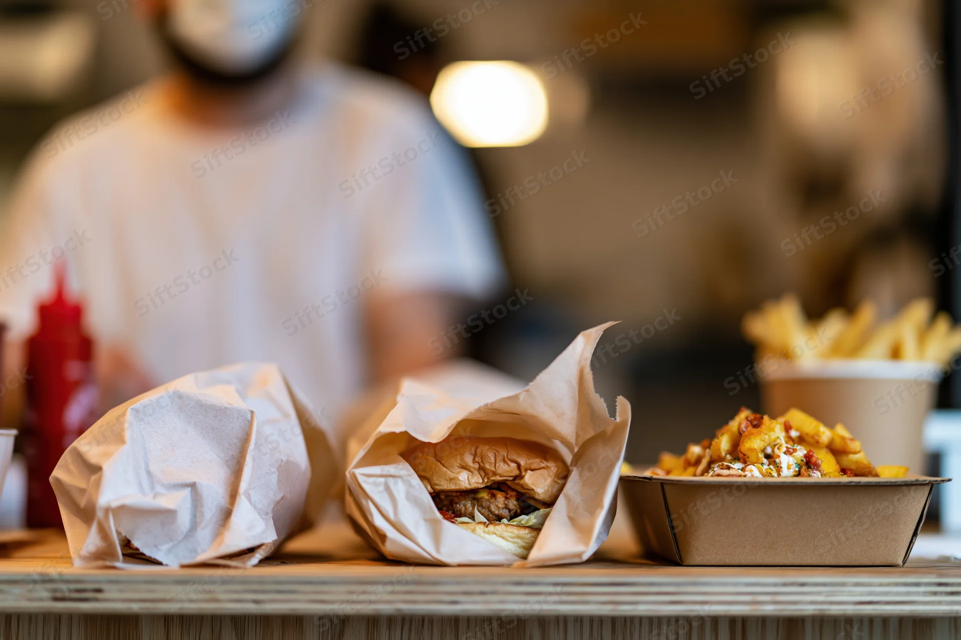 Two burgers wrapped in parchment paper and fries on a counter with a blurred worker in the background
