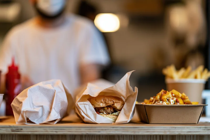 Two burgers wrapped in parchment paper and fries on a counter with a blurred worker in the background
