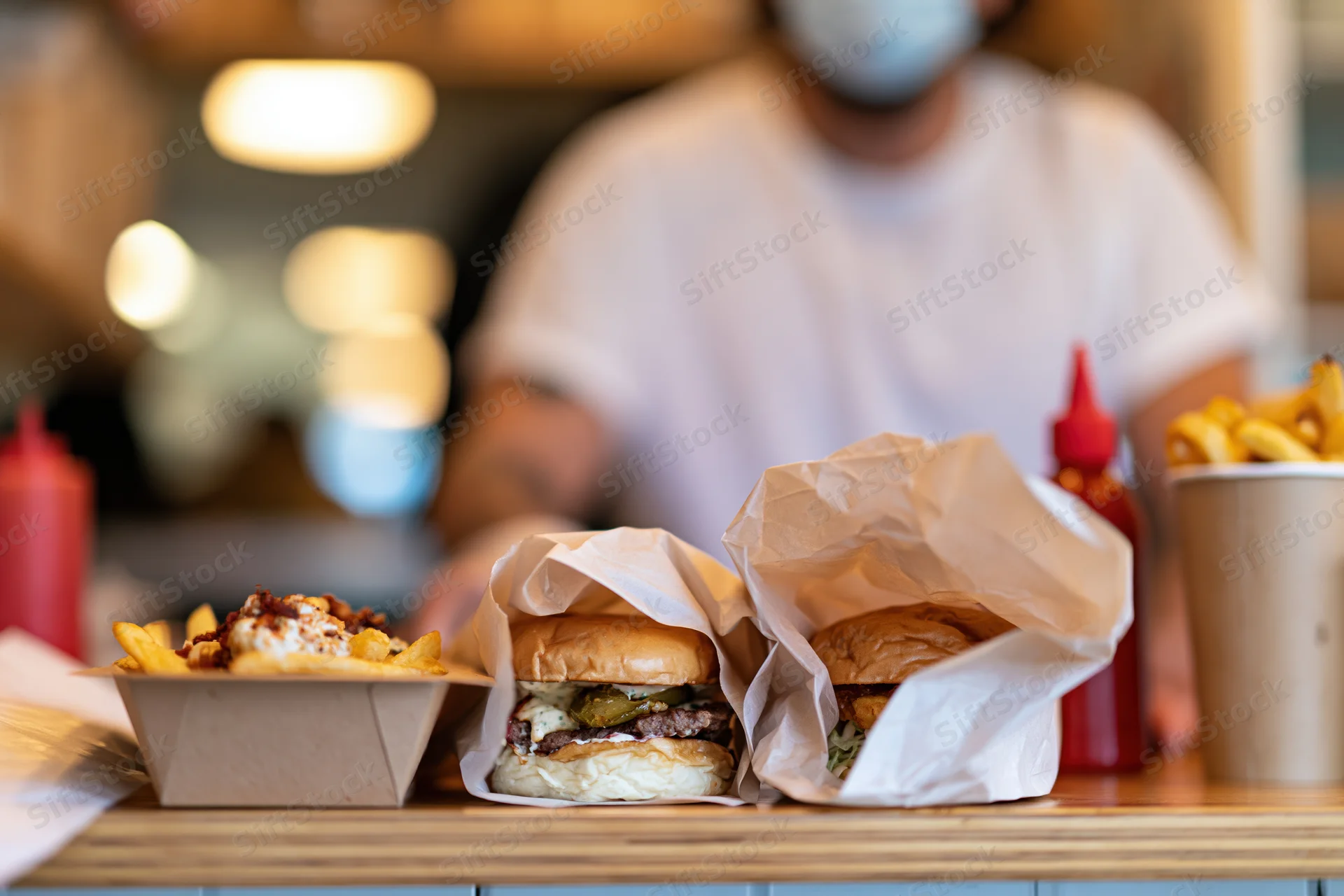 Two burgers wrapped in parchment paper and fries on a counter with a masked worker in the background