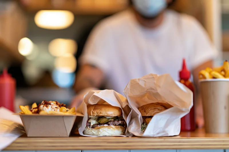 Two burgers wrapped in parchment paper and fries on a counter with a masked worker in the background