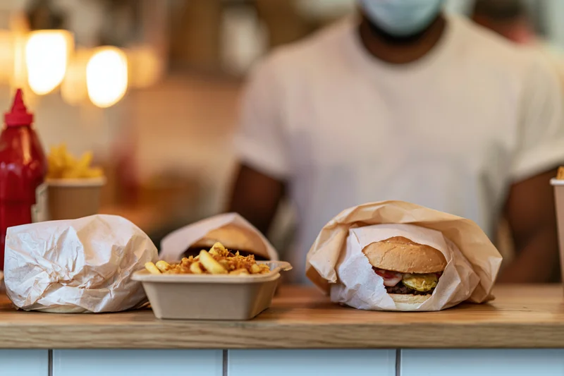 Two burgers wrapped in parchment paper and a container of fries on a wooden counter with a person in the background wearing a face mask