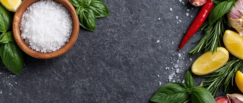 Coarse sea salt in a wooden bowl surrounded by fresh herbs and citrus on a dark slate surface