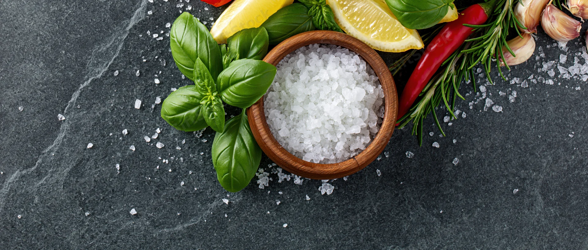 Coarse sea salt in a wooden bowl surrounded by fresh herbs and spices on a dark slate surface