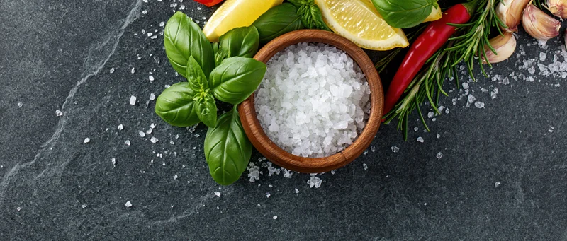 Coarse sea salt in a wooden bowl surrounded by fresh herbs and spices on a dark slate surface