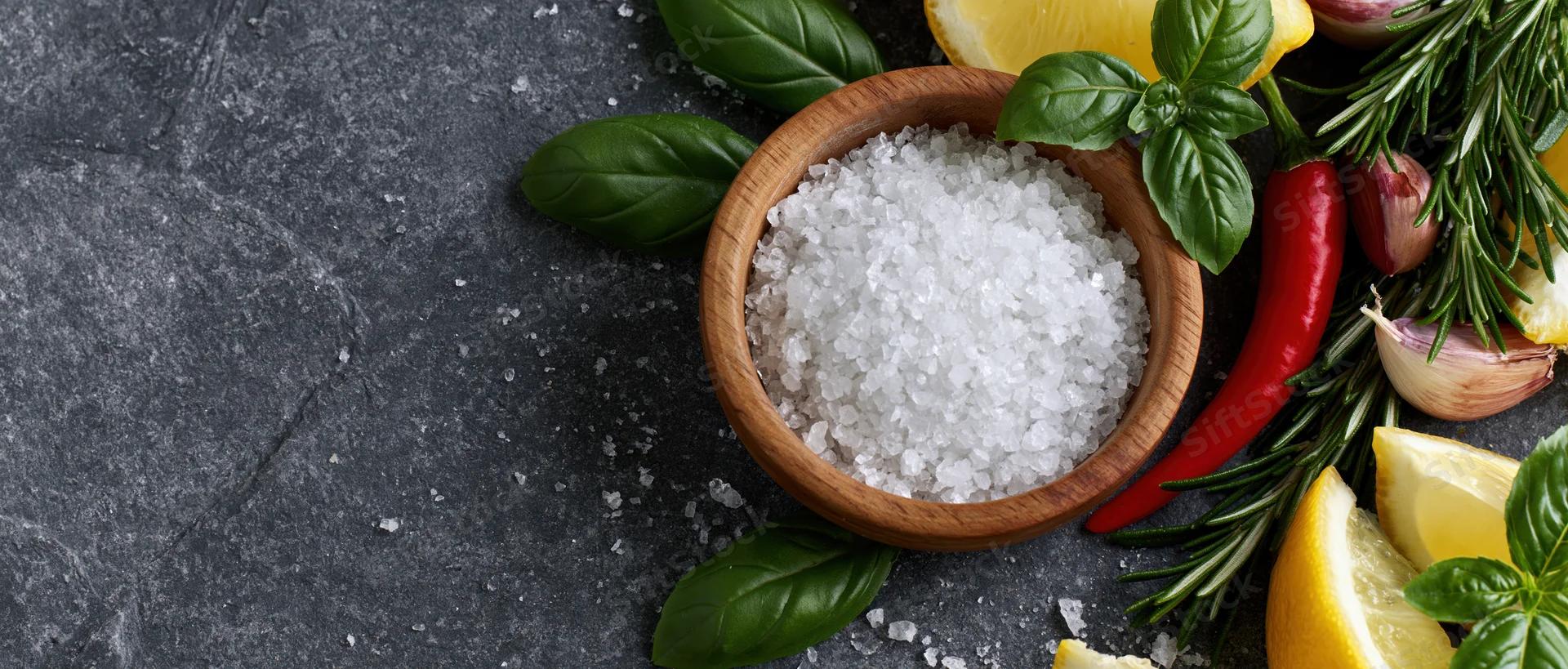Coarse sea salt in a wooden bowl surrounded by fresh herbs and citrus on a dark slate surface