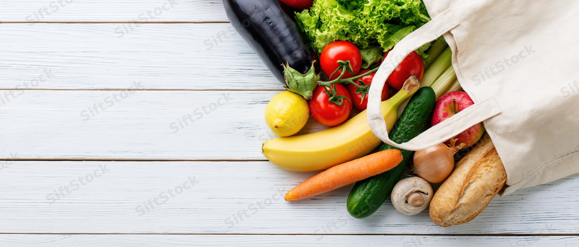 Cream canvas tote bag tipped on side spilling fresh vegetables and fruits onto white wooden table