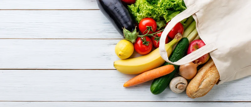 Cream canvas tote bag tipped on side spilling fresh vegetables and fruits onto white wooden table