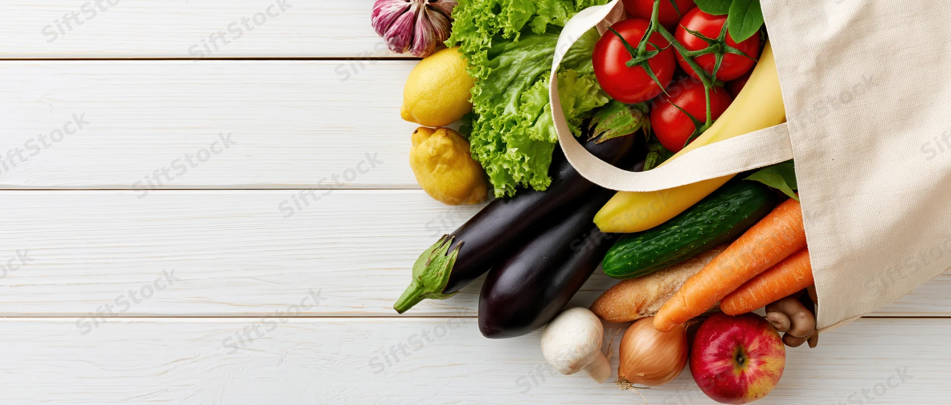 Cream canvas tote bag tipped on side spilling fresh vegetables and fruits on white wooden surface
