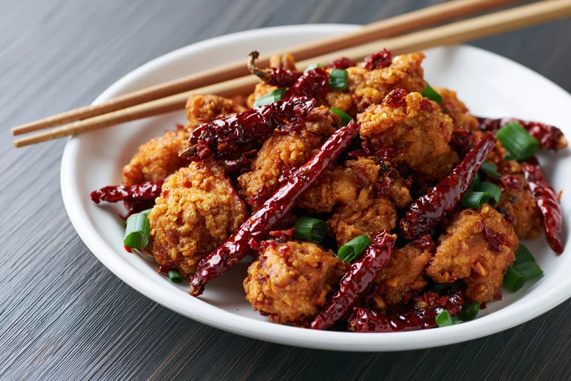 Crispy fried chicken pieces with dried red chilies and green onions on a white plate with chopsticks on a dark wooden table
