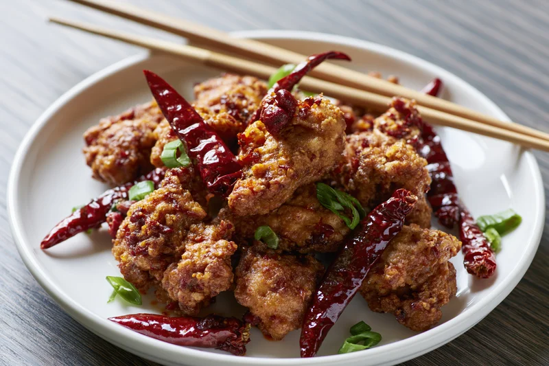 Crispy fried chicken pieces with dried red chilies and green onions on a white plate with chopsticks on a wooden table
