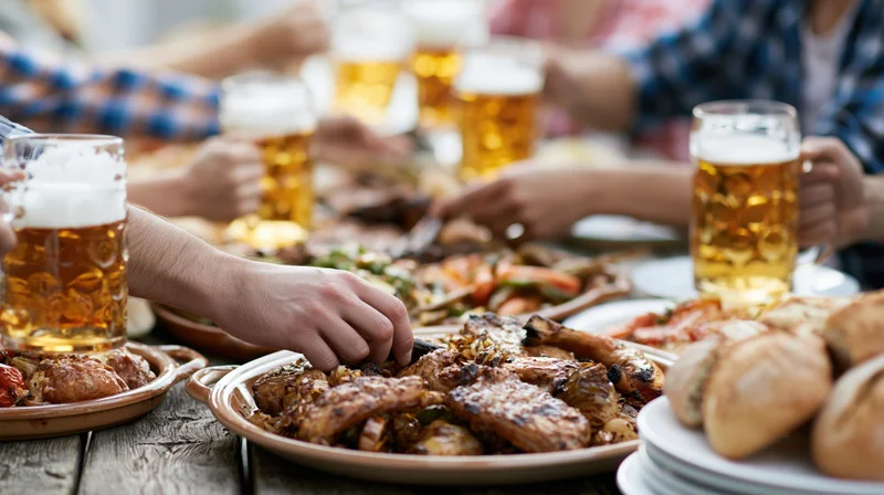 People sharing grilled meats and vegetables at a wooden table with beer glasses during an outdoor gathering