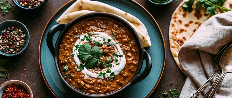 Dal makhani in a dark cast iron bowl topped with cream swirl, served with naan on a dark surface, surrounded by spices and garnishes, overhead view