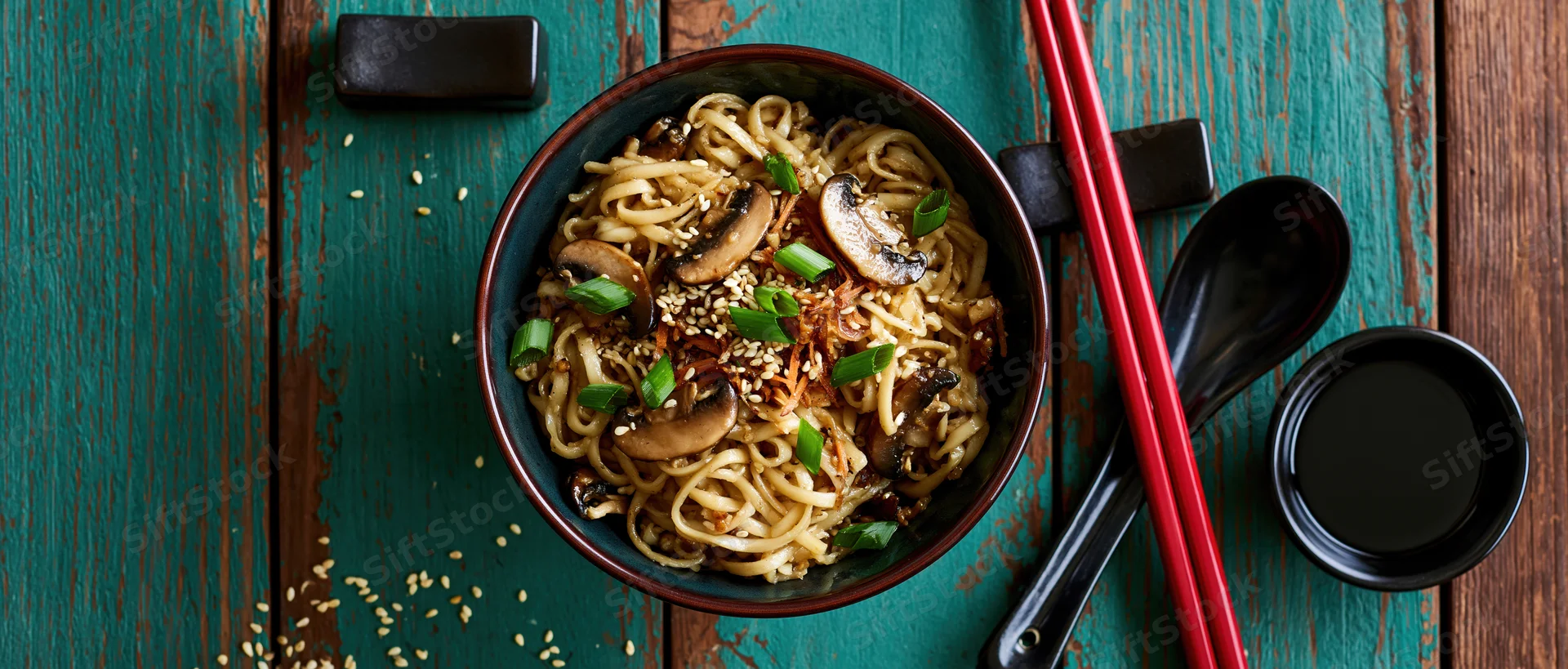 Bowl of ramen noodles with mushrooms and green onions on a teal wooden table, viewed from above with chopsticks and soy sauce nearby