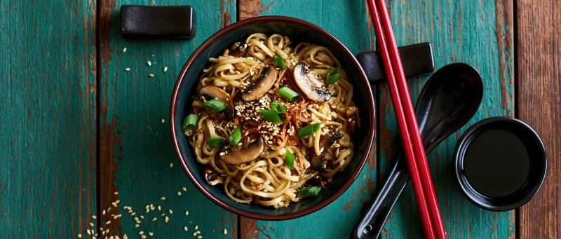 Bowl of ramen noodles with mushrooms and green onions on a teal wooden table, viewed from above with chopsticks and soy sauce nearby