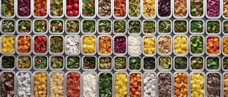 Dozens of aluminum foil takeout containers arranged in a grid, filled with colorful prepackaged meals and ingredients