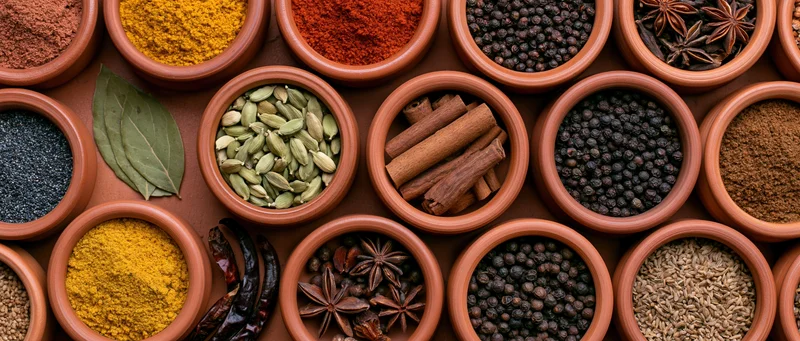 Overhead view of terracotta bowls filled with various whole and ground spices arranged in a grid pattern on a wooden surface