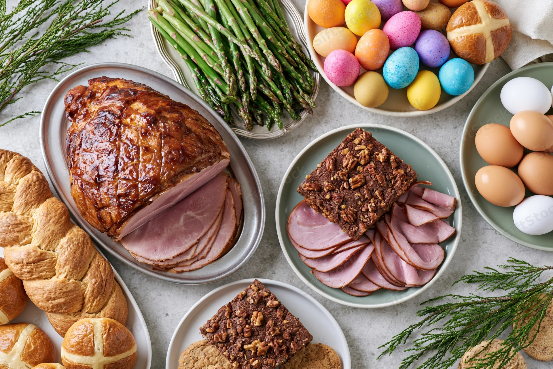 Easter holiday spread with ham, asparagus, colored eggs, hot cross buns and desserts arranged on a light grey stone surface