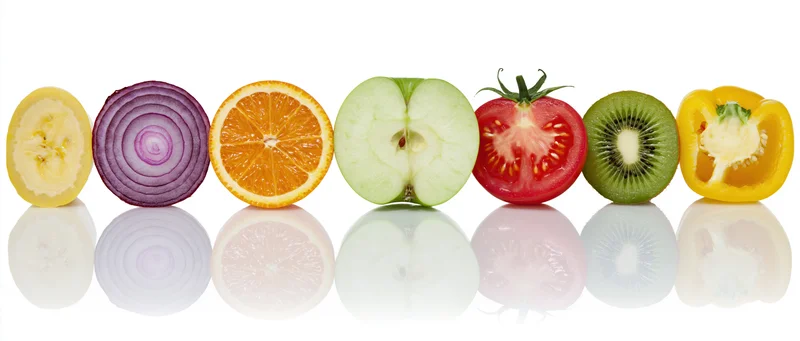 Eight fruit and vegetable cross-sections arranged in a row on a reflective white surface