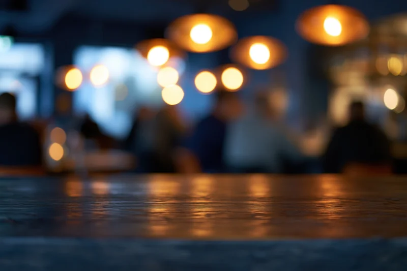 Empty dark wooden bar counter in foreground with blurred background of people and warm lights