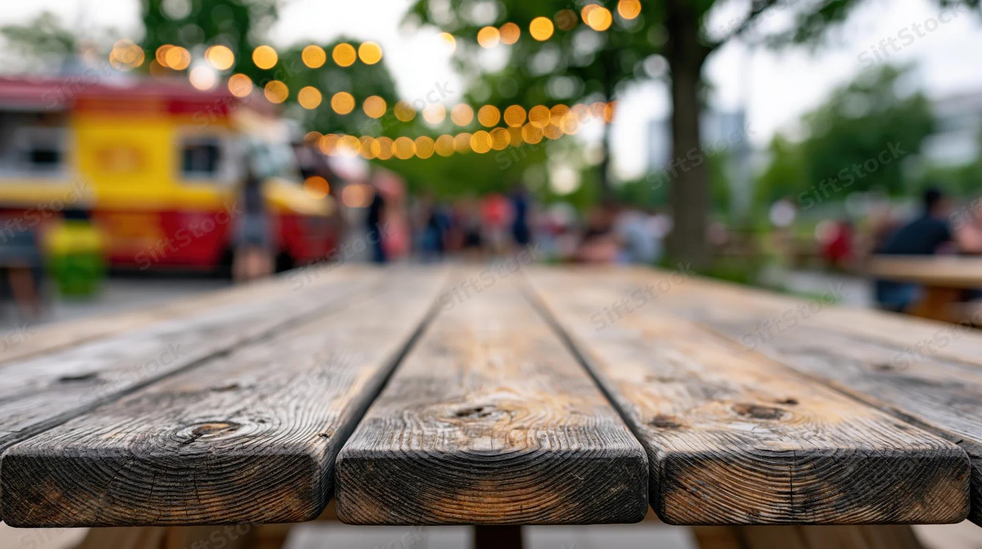 Worn wooden picnic table surface in sharp focus with blurred food truck and string lights in background