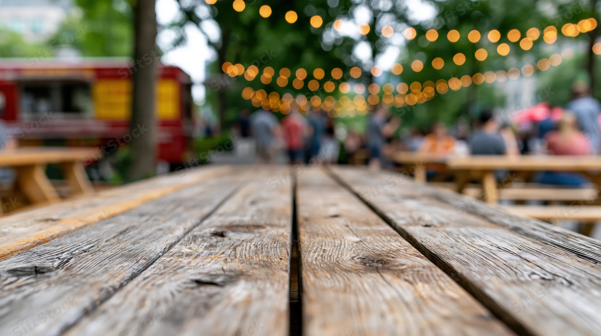 Worn wooden picnic table surface in sharp focus with blurred outdoor dining area and string lights in background