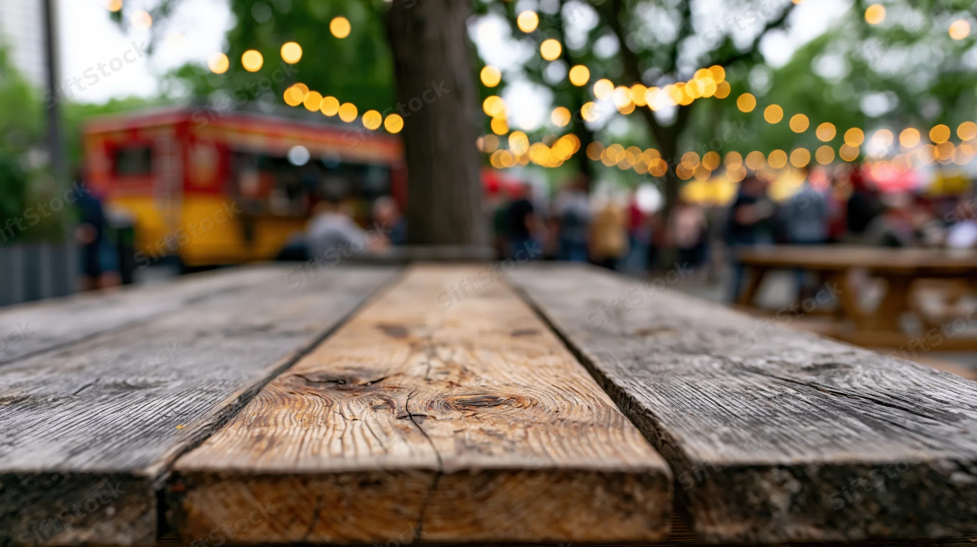 Worn wooden picnic table surface in sharp focus with blurred outdoor festival scene in background