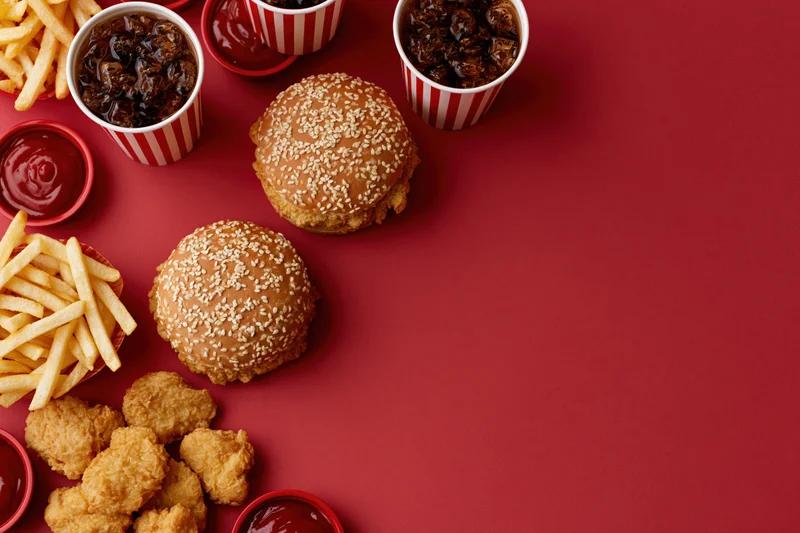 Fast food items including burgers, fries, chicken nuggets and ketchup arranged on a red surface in a flat lay composition