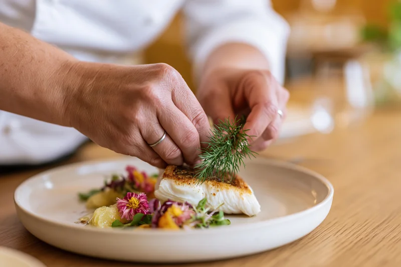 Chef's hands placing a sprig of dill on a seared fish dish on a plate