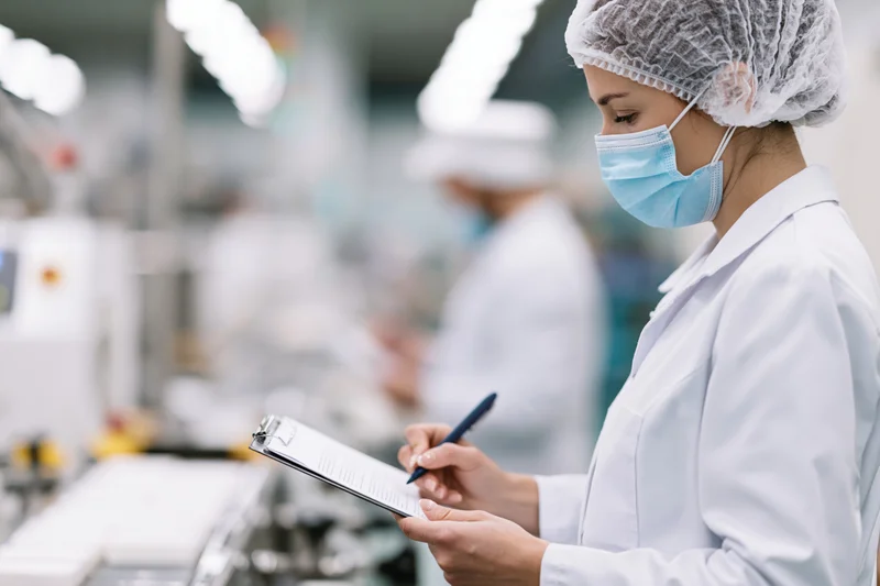 Woman in labcoat writing on clipboard during quality control inspection in cleanroom facility