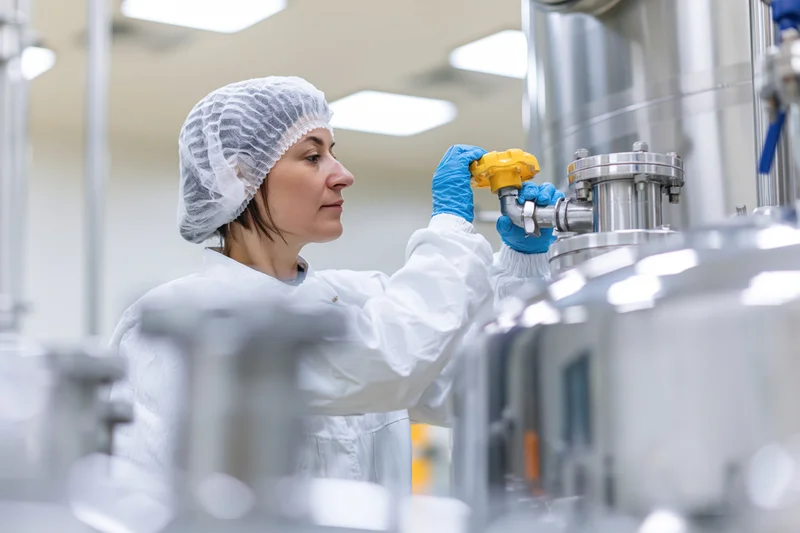 Woman in white protective coverall adjusting valve on stainless steel pipeline in cleanroom laboratory