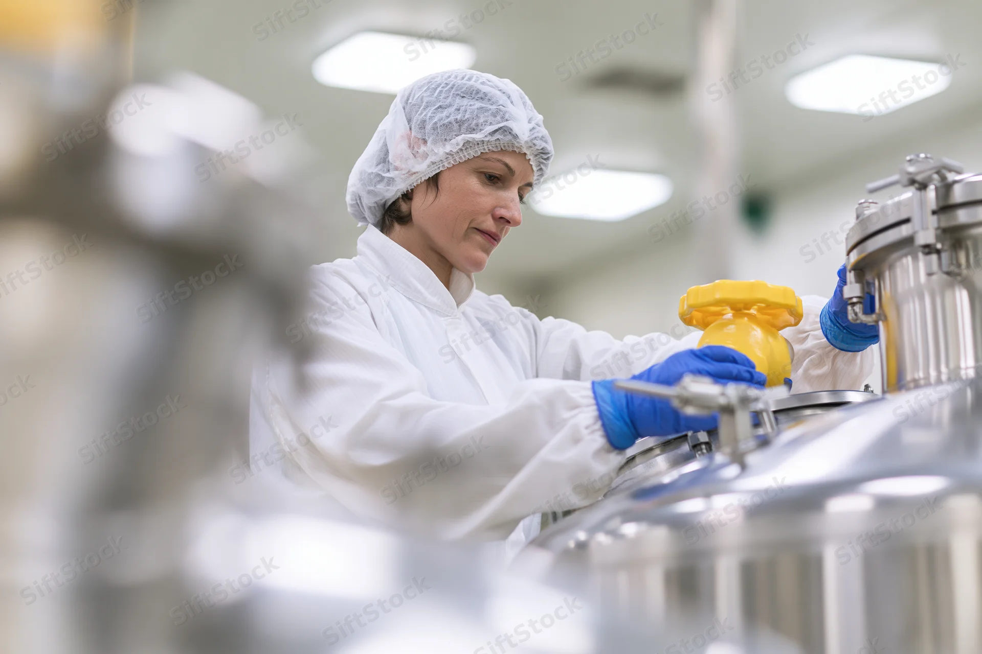 Woman in white coverall and hairnet adjusting valve on stainless steel tank in industrial facility