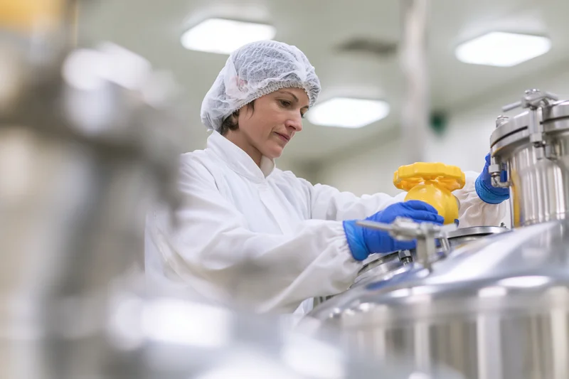 Woman in white coverall and hairnet adjusting valve on stainless steel tank in industrial facility