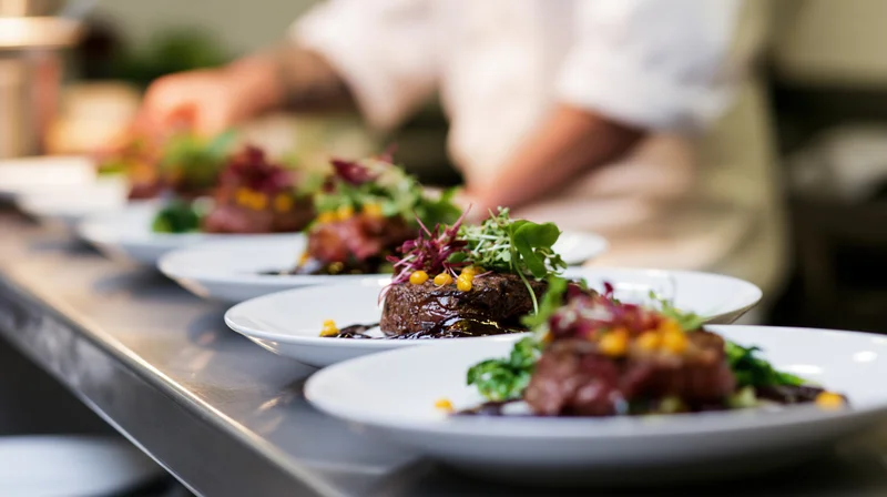 Fine dining plates arranged in a line on a stainless steel pass with a chef in the background