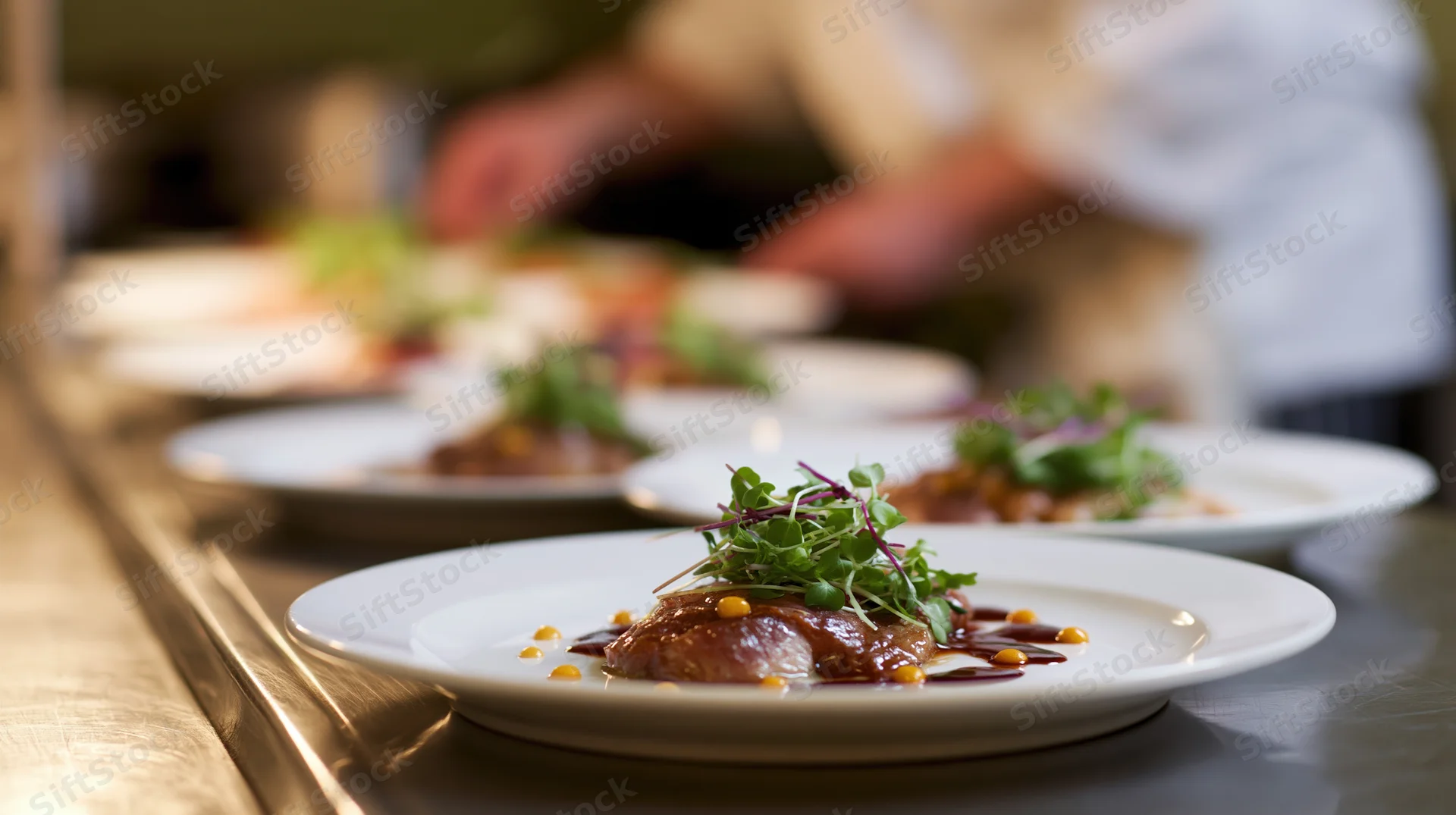 Fine dining plates arranged in a line on a stainless steel pass with garnished dishes in focus and chef in background