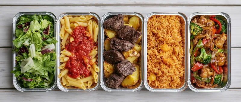 Five aluminum takeout containers in a row each filled with different meals including salad, pasta, beef with potatoes, rice, and stir-fry vegetables