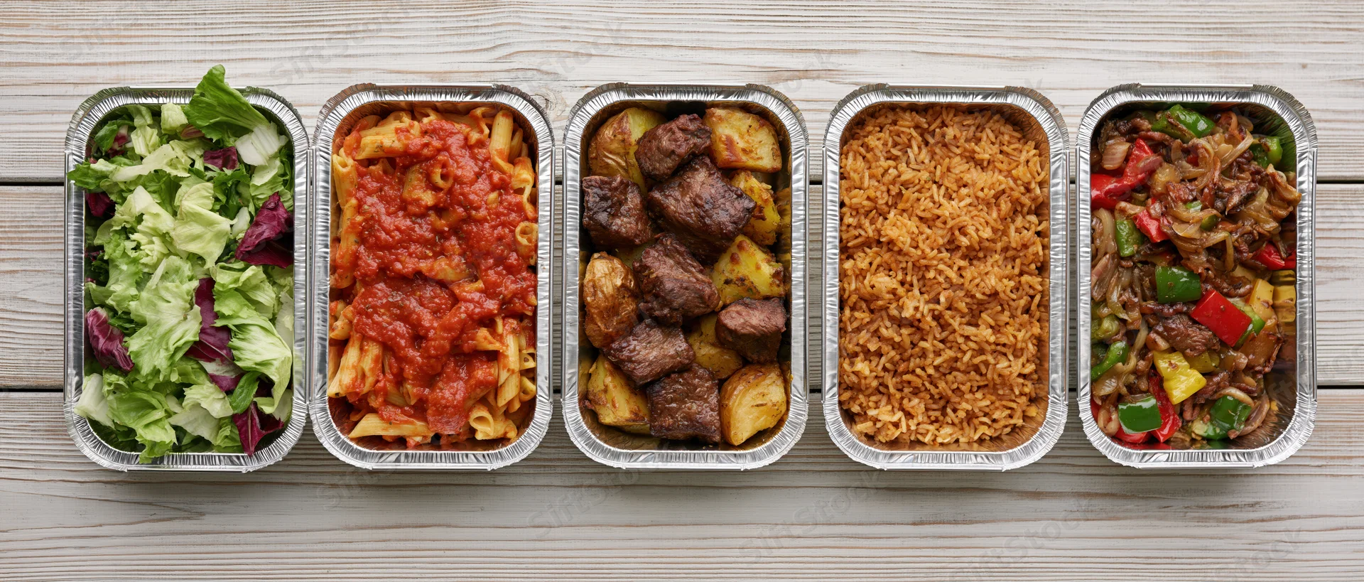 Five aluminum takeout containers in a row filled with different meals on a wooden surface