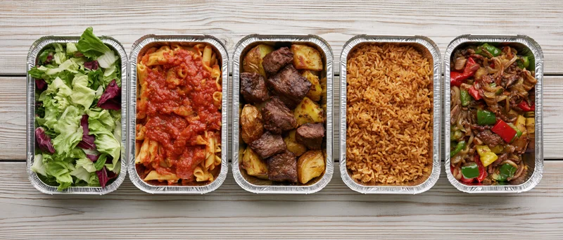 Five aluminum takeout containers in a row filled with different meals on a wooden surface