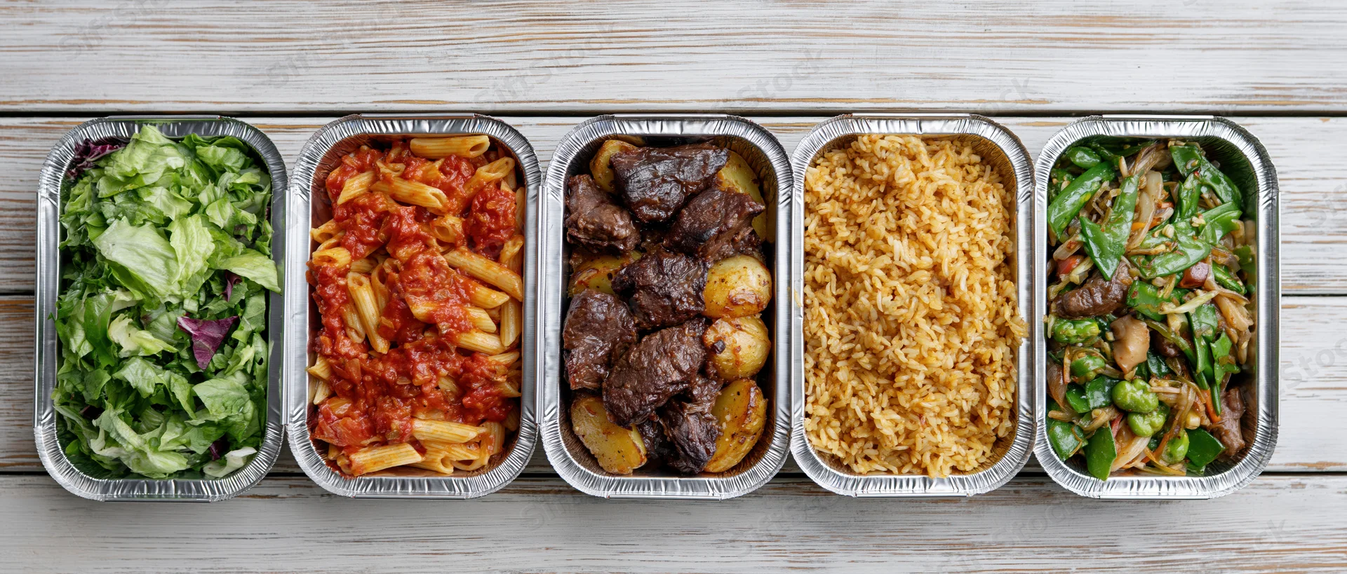 Five aluminum takeout containers in a row each filled with different meals on a wooden surface