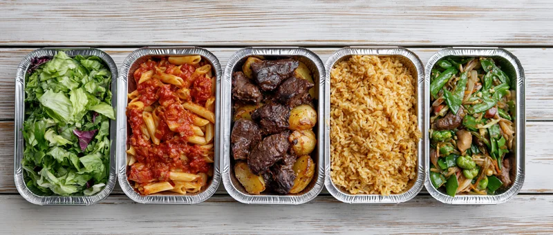 Five aluminum takeout containers in a row each filled with different meals on a wooden surface